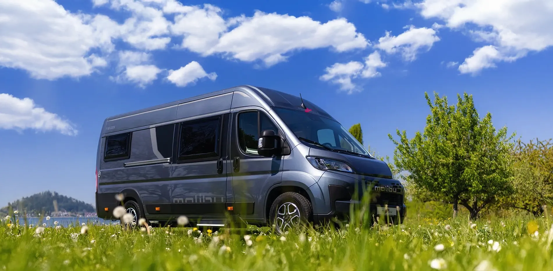 Graues Wohnmobil mit großen Fenstern auf einer Wiese unter blauem Himmel mit Wolken.